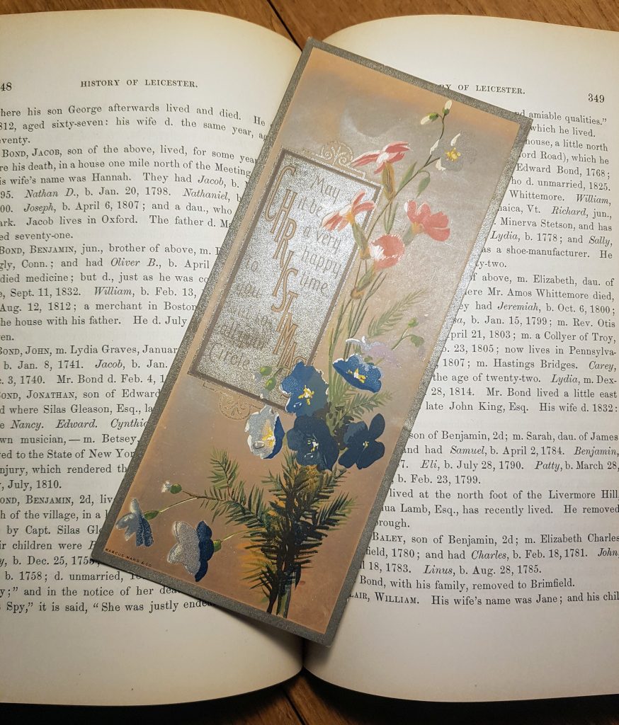 A victorian Christmas card placed on the pages of a book. Card reads "Christmas. May it be a very happy time to you and to all your circle"